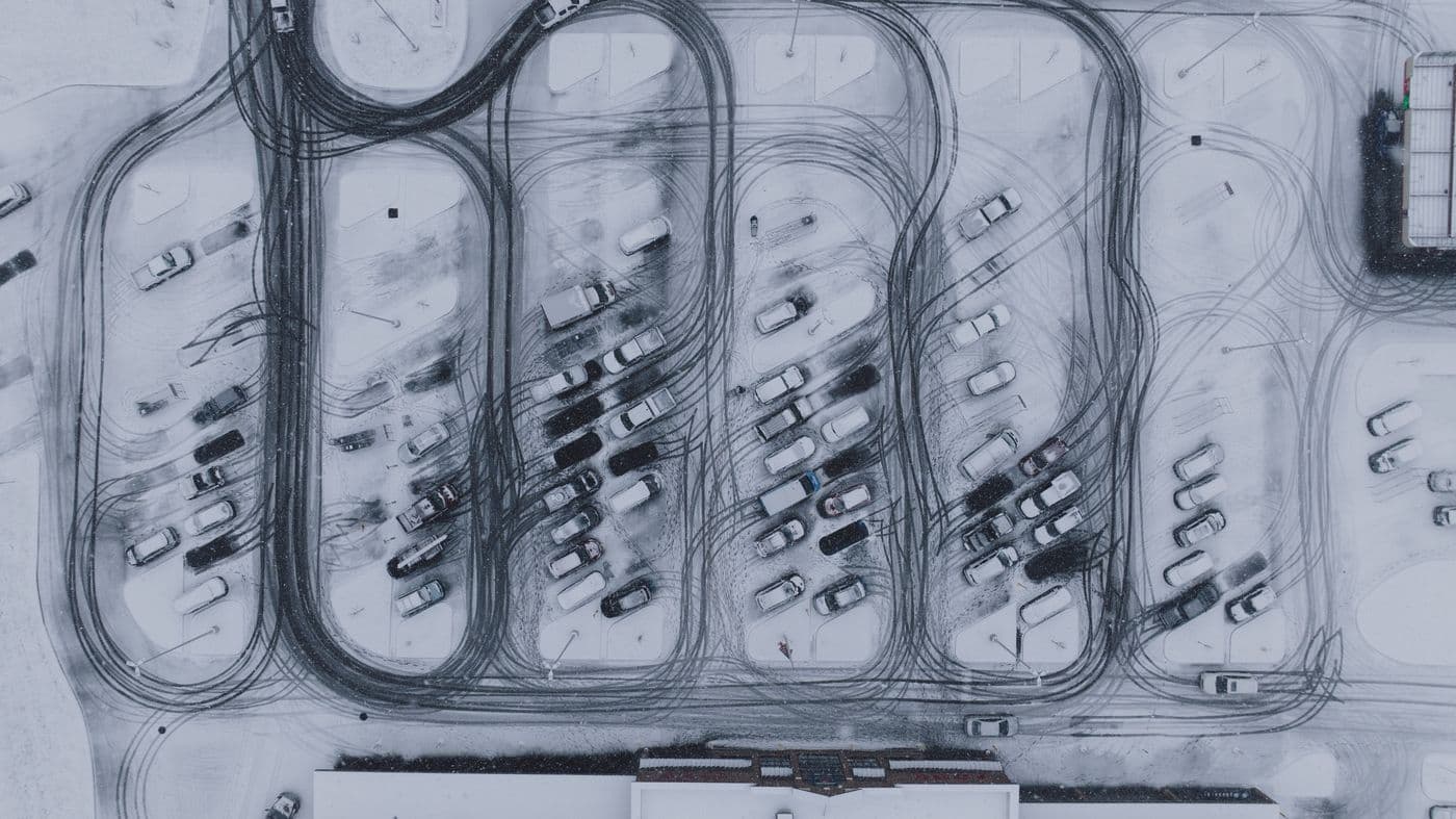 An aerial view of a plowed commercial parking lot with tire tracks through fresh snow.