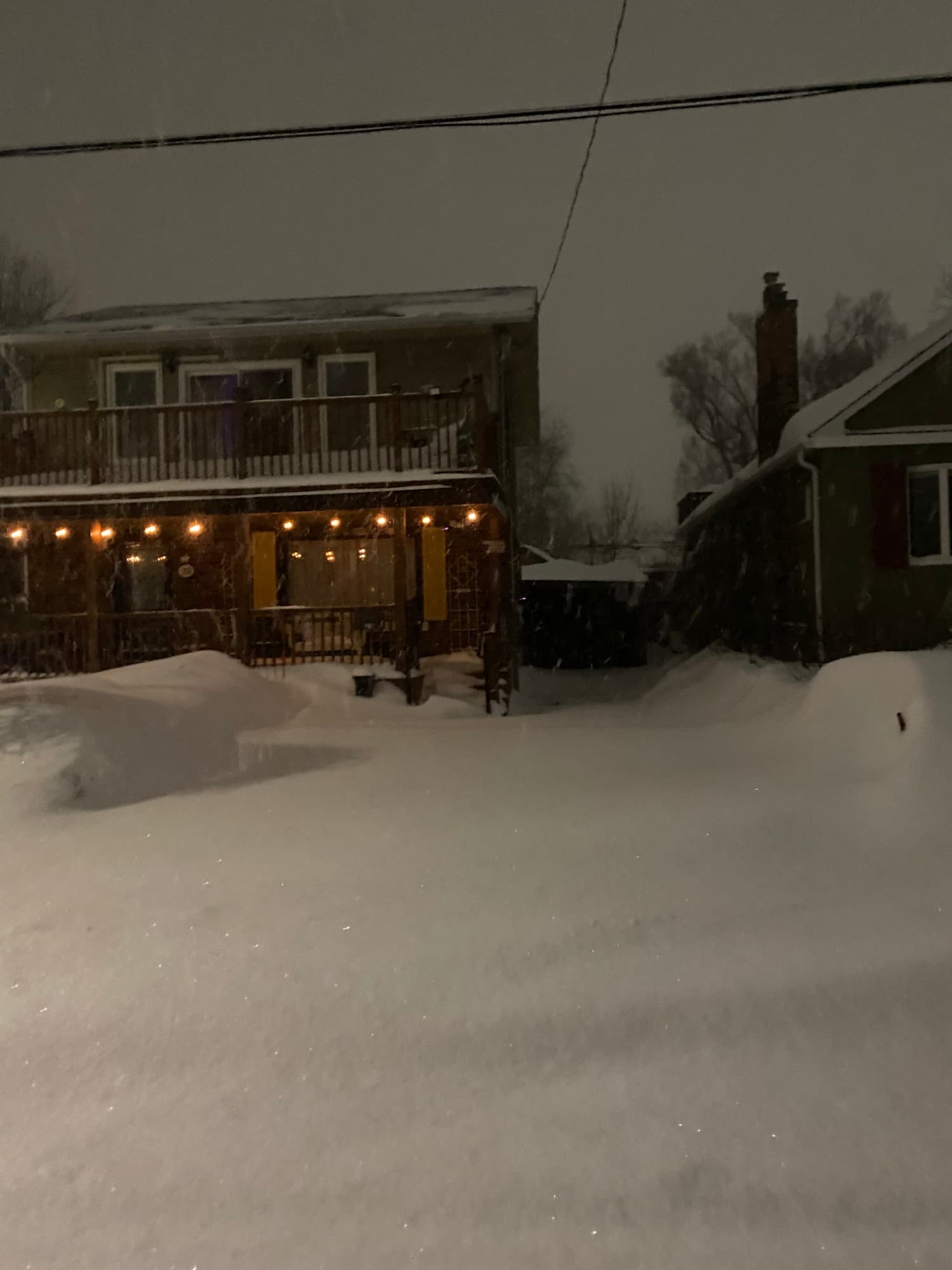 A two-storey house with string lights on the porch, the driveway buried under deep snow while more falls.