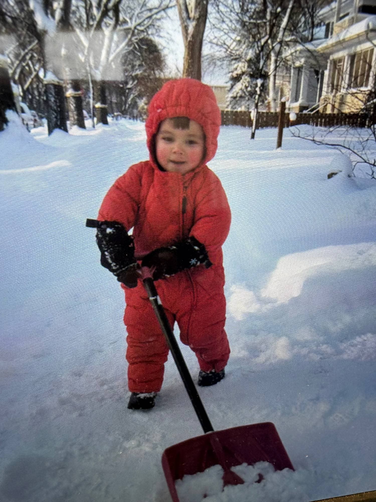 A toddler in a red snowsuit and black mittens pushing a red snow shovel across a snowy driveway.
