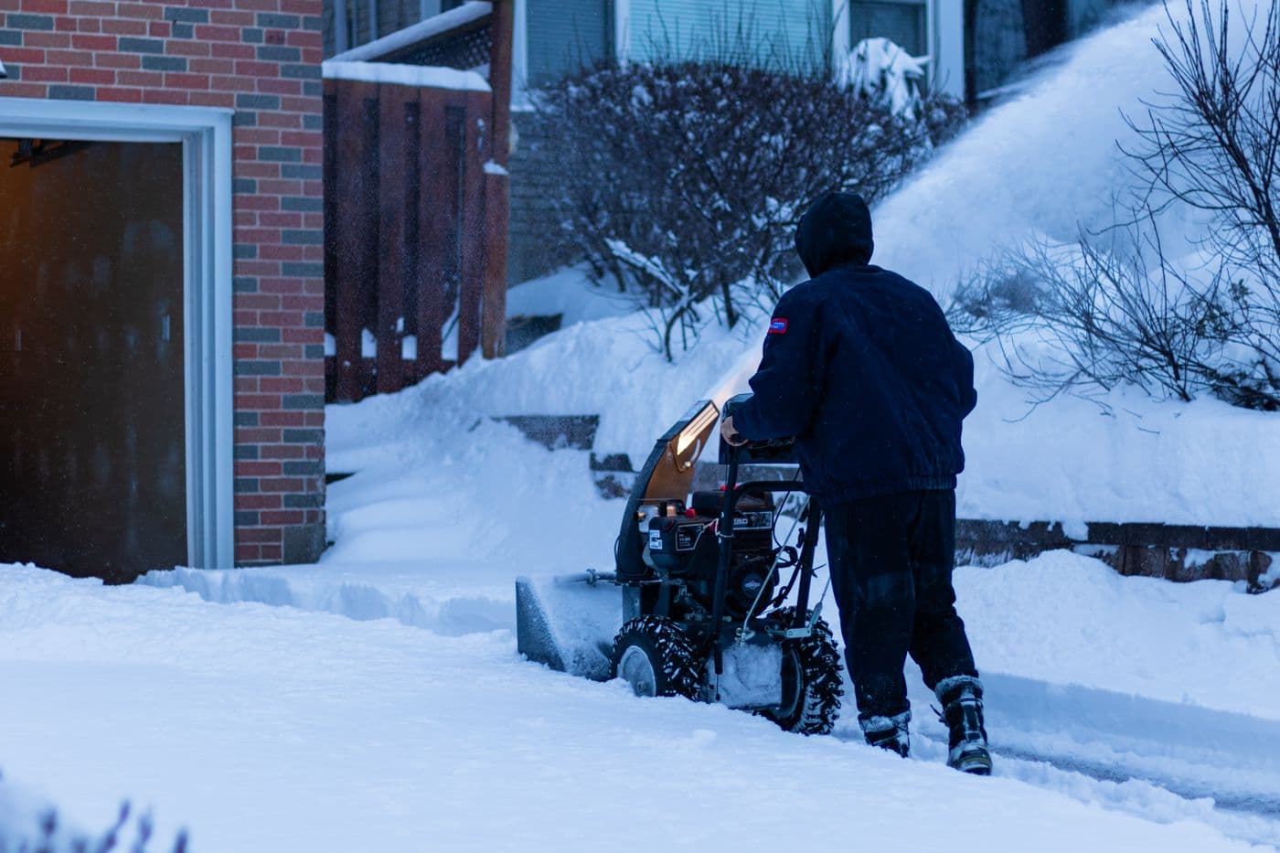 A snowblower throwing snow off a residential driveway, seen from behind.
